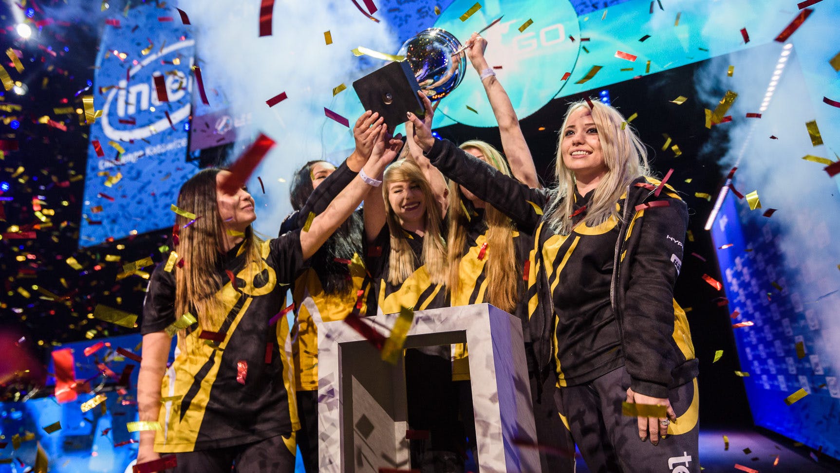 Een team van vrouwelijke e-sporters viert hun overwinning bij een esports-toernooi, omgeven door vallende confetti en juichend publiek.